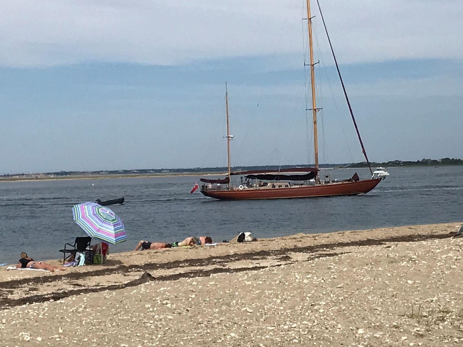 Beach goers watching sailboat in ocean.